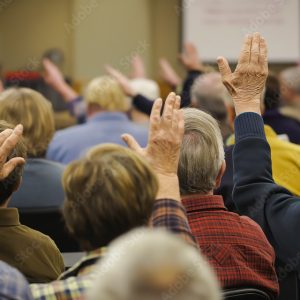 A politician addressing community issues during a town hall meeting with local residents raising hands for questions.