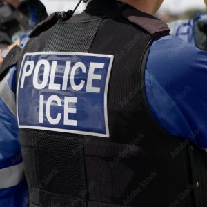 Ice Police Agent - officer of Immigration and Customs Enforcement. Close-up of POLICE ICE marking on the back of a stab proof vest worn by a trio of police officers at the scene of an incident.